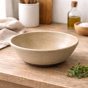 May include: A beige ceramic bowl with a dark brown base sits on a wooden surface. The bowl has a wide, shallow design. A sprig of green herbs is in the foreground. Other kitchen items are in the background.