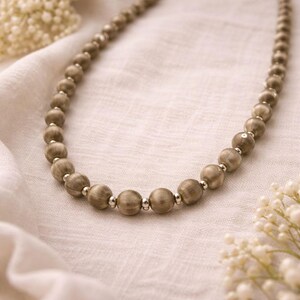 May include: A beaded necklace featuring large, round, light brown beads with silver-colored spacers. The necklace is displayed on a white, textured surface, with a soft focus background. The beads have a natural, organic appearance.