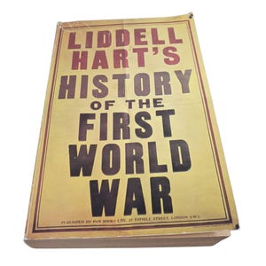 Puede incluir: Un libro antiguo titulado "Liddell Hart's History of the First World War". El libro tiene una cubierta amarilla con texto marrón y negro. El texto de la cubierta incluye el título y la información del editor.