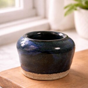 May include: A small, handcrafted ceramic pot with a dark blue and black glaze. The pot has a wide opening and a light beige base. The pot is sitting on a wooden surface, with a window and a plant in the background.