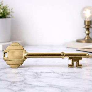 May include: A decorative brass key with a rectangular head and a detailed handle. The key is displayed horizontally on a marble surface. The key's design includes a rectangular opening and a decorative handle. The background includes a white wall and a small plant.