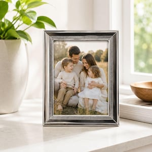 May include: A silver rectangular picture frame with a family portrait inside. The frame has a detailed border and is displayed on a white surface. The family in the photo includes two parents and two young children.