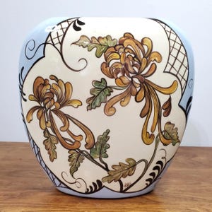 Puede incluir: Un jarrón de cerámica con borde azul claro y cuerpo crema. El jarrón presenta un diseño floral en tonos marrón, dorado y verde. El diseño incluye flores y hojas estilizadas. El jarrón tiene forma redondeada y está sobre una superficie de madera.