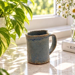 May include: A handmade ceramic mug with a textured, mottled blue glaze and an uneven, organic rim. The mug features a sturdy, thick handle and a cylindrical body with visible clay variations, showcasing a rustic, hand-thrown appearance.