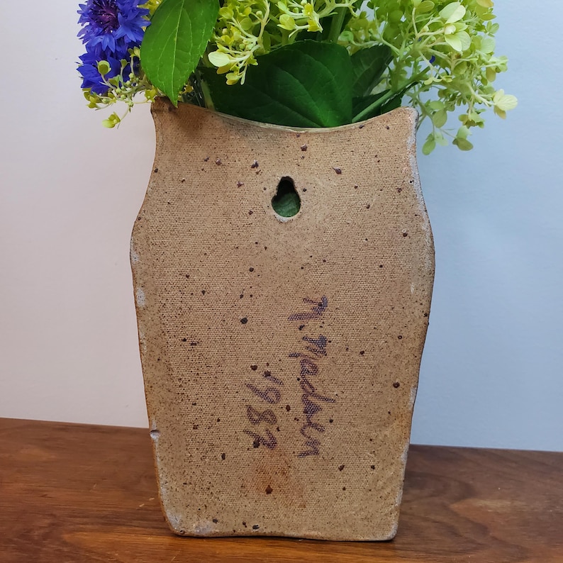 K&ouml;nnte beinhalten: Eine beige Wandtaschenvase aus Keramik mit strukturierter Oberfl&auml;che, die einen Strau&szlig; blauer und gr&uuml;ner Blumen enth&auml;lt. Die Vase hat ein Loch zum Aufh&auml;ngen und handschriftlichen Text mit der Aufschrift "M. Madsen 1983."