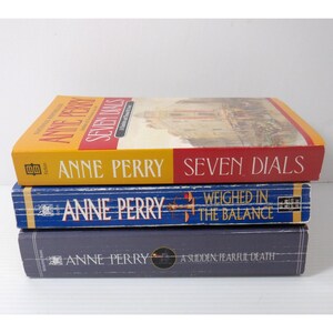 May include: Three hardcover books by Anne Perry are stacked. The top book is yellow and red, titled "Seven Dials." The middle book is blue, titled "Weighed in the Balance." The bottom book is dark blue, titled "A Sudden, Fearful Death."