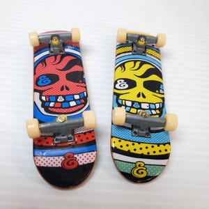May include: Two miniature skateboards with a skull design. One skateboard has a red skull design, and the other has a yellow skull design. Both skateboards have a black base with colorful stripes and the letter "&" on the bottom.