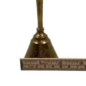 May include: A vintage brass bell with a conical shape and a long handle. The bell has etched designs and measures approximately 3 inches in height. A ruler is placed next to the bell for scale.