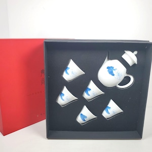 May include: A white porcelain tea set with blue floral designs, comprising a teapot and five small cups, presented in a black box. The set is packaged in a red box. The tea set is designed for serving tea.