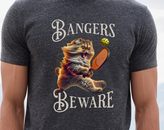 May include: A dark gray t-shirt with a white graphic of a cat holding a paddle and a yellow ball. The text "Bangers Beware" is written in a white cursive font.