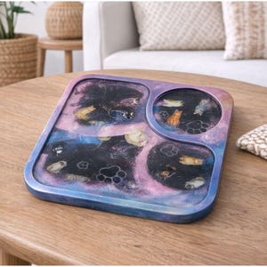 Pode incluir: Uma bandeja quadrada de resina com um design temático de galáxia. A bandeja tem um fundo escuro com elementos temáticos de gatos embutidos, incluindo pegadas e figuras de gatos. As bordas da bandeja são um gradiente de azul e roxo.