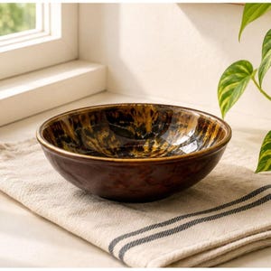 May include: A round ceramic bowl featuring a deep brown exterior and a glossy interior with a mottled, marbled pattern of dark brown, black, and golden yellow tones. The bowl has a smooth, curved rim and a wide, shallow basin shape.