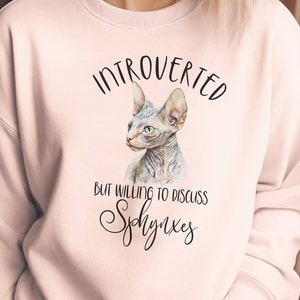 May include: Pink sweatshirt with a black and white illustration of a hairless cat. The text "Introverted but willing to discuss Sphynx" is printed on the sweatshirt.