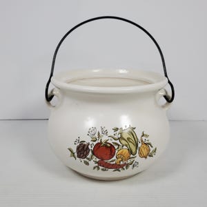 Puede incluir: Olla de cerámica blanquecina con asa negra e ilustraciones de verduras. La olla tiene forma redonda con una abertura ancha y dos asas pequeñas. Las ilustraciones de verduras incluyen un tomate, pimiento y champiñón.