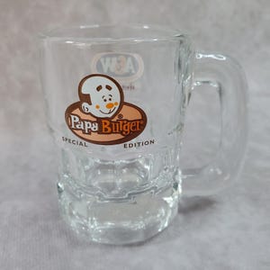 May include: Clear glass mug with a handle, featuring a Papa Burger logo in brown and white. The logo includes a cartoon character and the words "Special Edition". The mug has a rectangular base.