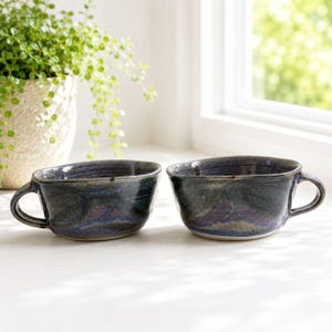 May include: Two wide, low-profile ceramic mugs with a dark, mottled glaze featuring deep blue, purple, and black tones. Each mug has a single curved handle and a slightly irregular, organic rim shape, showcasing a glossy, reflective finish.