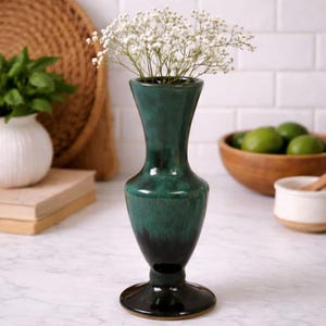 May include: A dark green ceramic vase with a flared top and a wide base, holding a bouquet of small white flowers. The vase has a glossy finish and is displayed on a white countertop. Other items in the background include a white vase, a wooden bowl, and a woven basket.