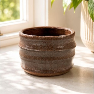 Op de afbeelding: Een bruine keramische plantenpot met een textuur, geribbeld ontwerp. De pot heeft een brede opening en een donker interieur. De buitenkant heeft een rustieke, aardse uitstraling, geschikt voor binnen of buiten. De pot staat op een wit oppervlak.