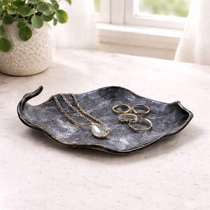 Op de afbeelding: Een donkergrijze, bladvormige sieradenbak bevat een gouden ketting met een druppelvormige hanger en verschillende gouden ringen. De bak heeft een getextureerd oppervlak en een gebogen handvat.