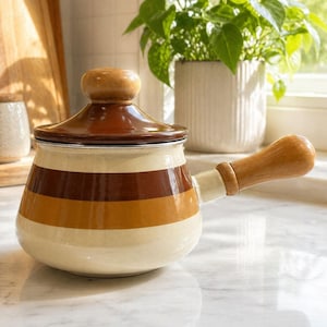 May include: A vintage enamel saucepan with a wooden handle and lid. The pot has cream, brown, and orange horizontal stripes. The lid features a wooden knob. The saucepan is on a marble surface, with a plant in the background.