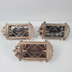 Pode incluir: Três aldravas de porta antigas em metal tom bronze. Cada aldrava tem uma moldura retangular com um design ornamental e enrolado e uma placa traseira decorativa. As aldravas estão dispostas sobre uma superfície branca.