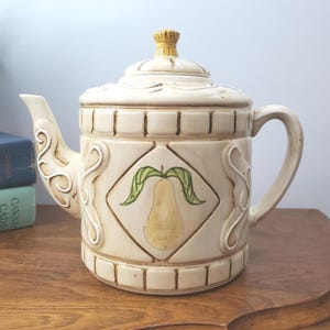 May include: Cream ceramic teapot with a lid and handle. The teapot has a pear design on the front and decorative scrollwork. The lid has a textured, straw-like knob. The teapot is on a wooden surface.