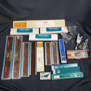 Könnte beinhalten: Eine Sammlung von Zeichenwerkzeugen und -zubehör, darunter Radiergummis, technische Stifte und Zeichenvorlagen. Die Artikel befinden sich in verschiedenen Schachteln und Etuis, mit Marken wie Faber-Castell und Pelikan. Die Werkzeuge sind auf einer dunklen Oberfläche angeordnet.