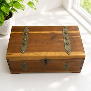 May include: A rectangular wooden box featuring two vertical brass-toned metal straps with decorative rivets on the lid and two smaller matching plates on the front. The box has a central metal latch and visible finger-jointed corner construction.