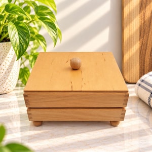 May include: A square wooden box with a flat lid and a small round wooden knob handle. The box features visible finger joint construction on the corners and rests on four small, rounded wooden feet, showcasing a natural light brown wood grain.
