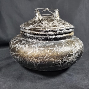 Puede incluir: Un tarro decorativo con tapa, base negra y dorada, adornado con vetas blancas en forma de telaraña. La tapa tiene un asa con un detalle de alambre. El tarro tiene una forma redondeada, adecuado para almacenamiento o exhibición.