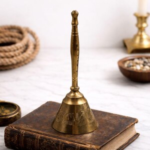 May include: A vintage brass bell with a long handle and a flared base, resting on a worn, leather-bound book. The bell features etched designs and has a warm, golden hue. A nautical theme is suggested by the rope in the background.
