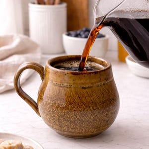May include: A brown ceramic mug is being filled with dark coffee from a glass carafe. The mug has a textured, crackled surface and a large handle. The background includes white ceramic containers and a bowl of blueberries.