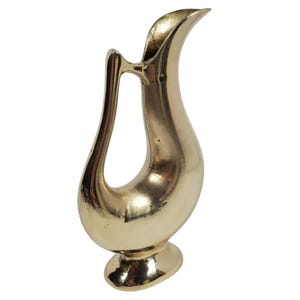 Puede incluir: Una jarra decorativa dorada con un asa curva y un pico acampanado. La jarra tiene un cuerpo bulboso que se estrecha hasta una base de pedestal. La superficie es lisa y reflectante, con un acabado pulido.
