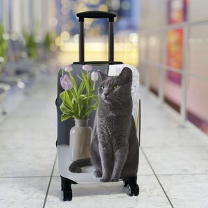 May include: A gray cat with green eyes sits in front of a vase of pink tulips on a suitcase with a black handle.