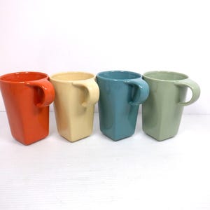 May include: Four square mugs with curved handles, in orange, cream, teal, and green. The mugs have a glossy finish and are set against a white background. Each mug is a unique colour.
