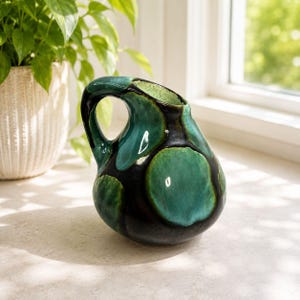 May include: A ceramic jug with a distinctive handle and a glossy finish. The jug has a dark teal and black colour scheme with large teal circular patterns. The jug is on a light-coloured surface near a window.
