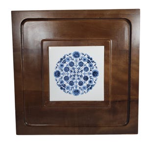 May include: A square wooden tray with a recessed border. The centre features a white tile with a blue floral design. The tray is a warm brown colour, and the tile has a detailed, circular pattern.