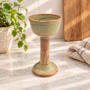 Puede incluir: Un cáliz de cerámica hecho a mano con un cuenco ancho y poco profundo de esmalte verde pálido y un tallo cilíndrico texturizado de color marrón rojizo. La base es circular y se ensancha, combinando con los tonos terrosos, verdes y marrones.