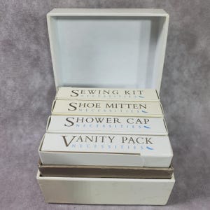 Op de afbeelding: Een open witte doos met vier kleinere dozen met de labels "Sewing Kit", "Shoe Mitten", "Shower Cap" en "Vanity Pack". Elke doos heeft het woord "Necessities" onder het hoofdlabel gedrukt. De dozen zijn gestapeld in de grotere doos.