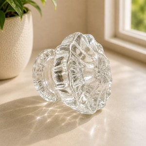 Puede incluir: Un pomo de cristal transparente para armarios con un diseño de bordes festoneados y múltiples facetas con una base circular. El material transparente refleja la luz, creando patrones intrincados en la superficie.