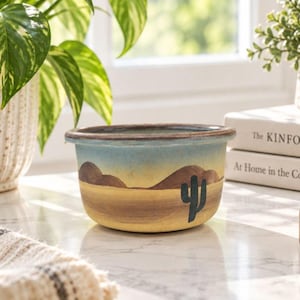 May include: A round ceramic bowl featuring a painted desert landscape with a blue sky, brown hills, and a yellow sandy foreground. A dark green saguaro cactus is painted on the side. The bowl has a wide, dark brown rim and a smooth, curved body.