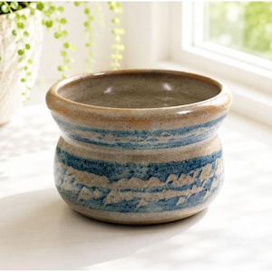May include: A round ceramic bowl with a wide, flared rim. The exterior features a speckled beige base color with horizontal bands of textured blue glaze. The interior of the bowl is smooth and finished in a light, neutral, speckled beige tone.