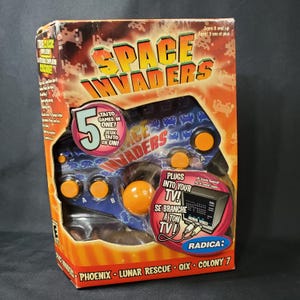 Peut inclure: Une console de jeu vidéo Space Invaders en boîte. La boîte est orange et porte le titre du jeu en grandes lettres. La console a des boutons orange et se branche sur un téléviseur. La boîte indique "5 jeux Taito en un!"