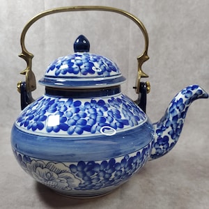 May include: A blue and white ceramic teapot with a floral design. It has a brass handle and spout, and a lid with a blue knob. The teapot's body is decorated with blue floral patterns on a white base, a classic design.