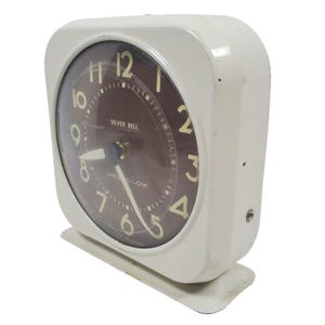 Può includere: Una sveglia Silver Bell vintage bianca con quadrante marrone e numeri dorati. L'orologio ha una forma rettangolare con bordi arrotondati e una piccola base. Il quadrante recita "Silver Bell" e "Westclox".