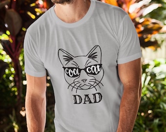 May include: A light gray t-shirt featuring a black graphic of a cat wearing sunglasses with the words "cool cat" on the glasses and the word "DAD" below. The person in the image is wearing sunglasses.