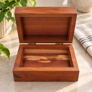 Peut inclure: Une boîte rectangulaire en bois avec un couvercle à charnières en position ouverte. La boîte présente une finition en grain de bois brun rougeâtre chaud et est construite avec des charnières métalliques visibles reliant le couvercle à la base.