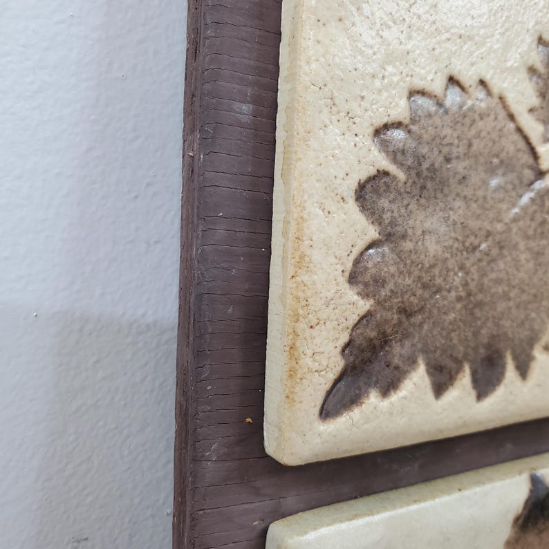 Puede incluir: Primer plano de una pieza de arte mural decorativa. La obra presenta baldosas de cer&aacute;mica cuadradas con un dise&ntilde;o de hojas en tonos marrones y beige. Las baldosas est&aacute;n colocadas dentro de un marco de madera marr&oacute;n oscuro, creando una est&eacute;tica r&uacute;stica.