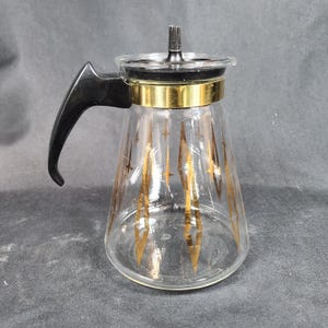 Può includere: Una caffettiera in vetro trasparente con manico e coperchio neri. La caffettiera ha una fascia dorata al centro e disegni dorati a stella. Stile vintage.