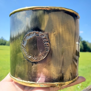 May include: A cylindrical container with a metallic finish. The exterior has a dark, textured base with gold accents and circular embellishments. The rim and base are trimmed with gold. The container is held against a backdrop of a green field and blue sky.